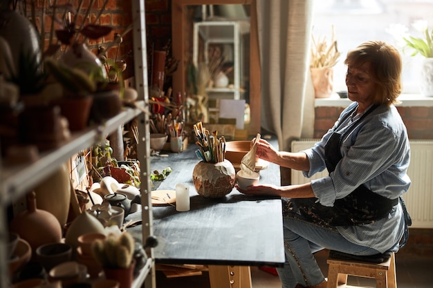 Nice female potter in apron holding bowl and paint brush while sitting at the table in pottery studio