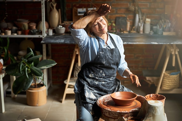 Nice female potter in apron wiping forehead with hand and smiling while working on pottery wheel