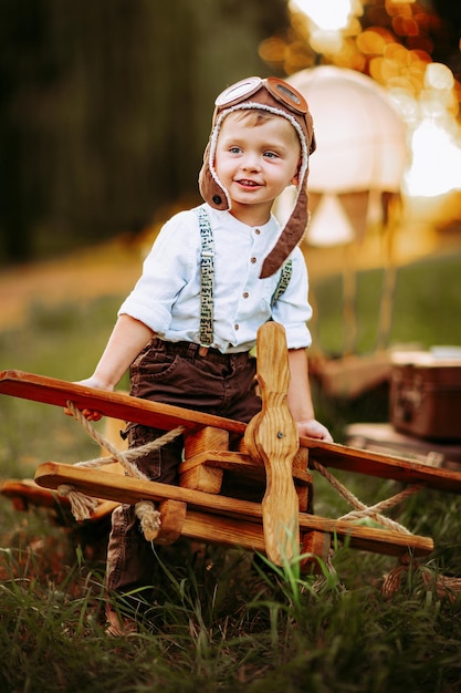 Nice little pilot boy in vintage aviator hat sitting on big toy wooden plane outdoor and playing