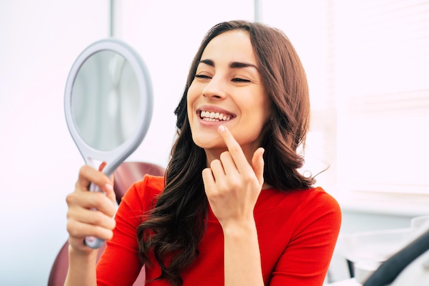 Nice looking girl in scarlet sweater is holding a mirror in her elegant hand and looking through the perfect result of the dental work