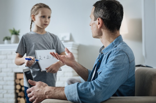 Nice lovely fair-haired little girl holding a notebook and standing near her daddy and asking his help with math while he working