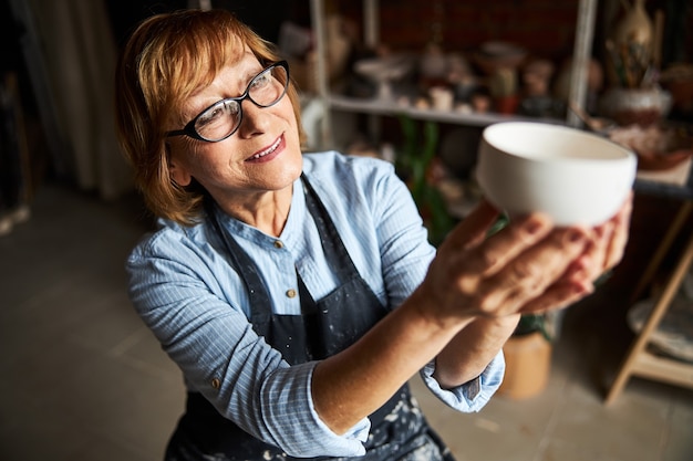 Nice old woman ceramic artist holding handmade earthenware bowl and smiling while working in pottery studio