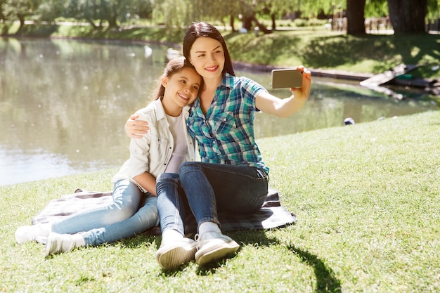 Nice picture of mom and daughter taking selfie. They are sitting on blanket near pond. Woman is holding phone. They are looking at it and smiling. Girls are posing.