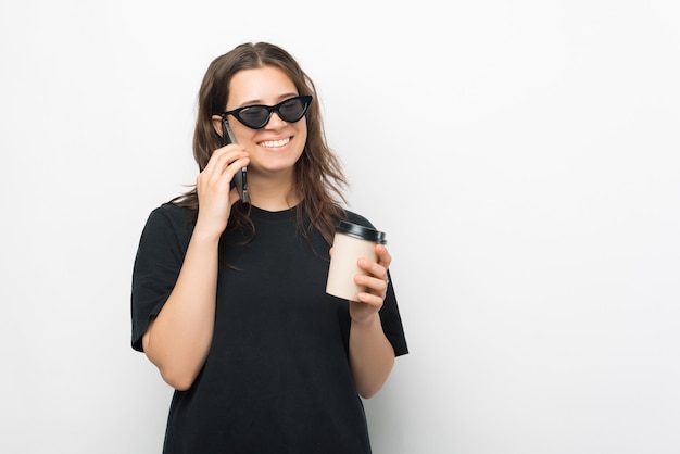 A nice picture of a young woman drinking her hot drink while talking on the phone smiling with sun glasses on