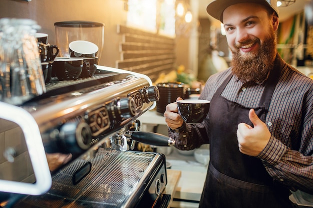 Nice and positive young barusta stand in kitchen at coffee machine. He hold big thumb up and cup of coffee. Guy smiles to camera.