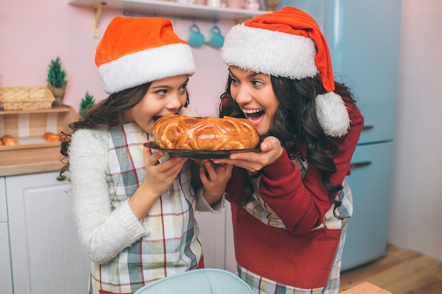 Nice and positive young woman and girl hold plate with pie and look at each other. They bite it. Woman and girl smile. They wear aprons and Christmas hats.