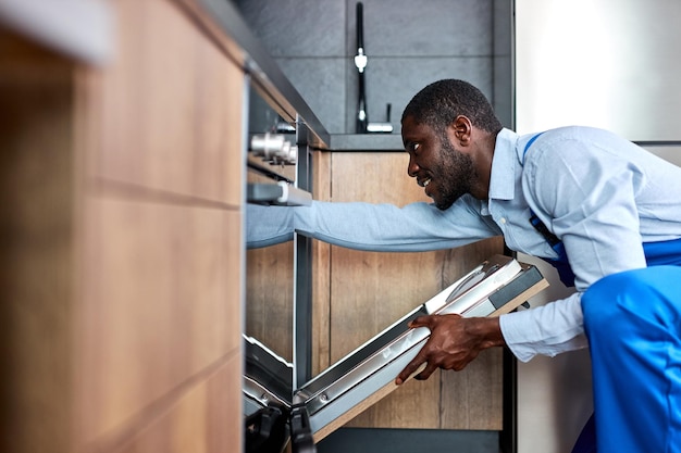 Nice professional african american handyman in blue overalls uniform is going to repair dishwasher c...