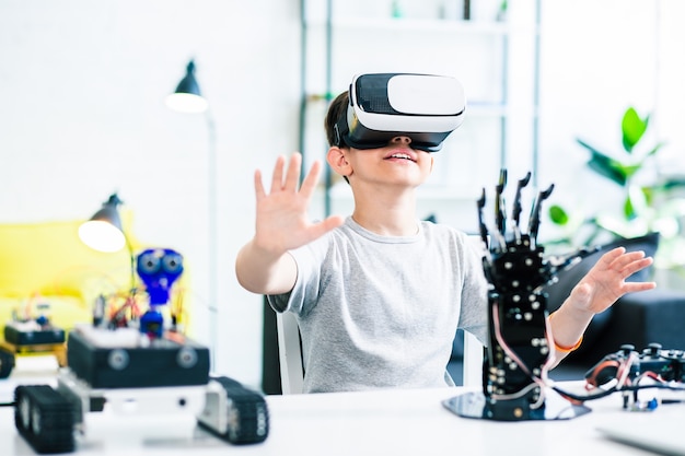 Nice smart enthusiastic boy sitting at the table and wearing VR glasses while experimenting with his robotic devices