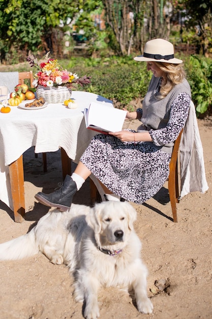 A nice woman in a hat is sitting near a table on the street next to a white longhaired labrador