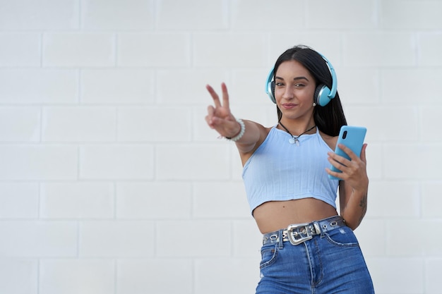 Nice young girl making the victory sign while listening to music with her smartphone and headphones