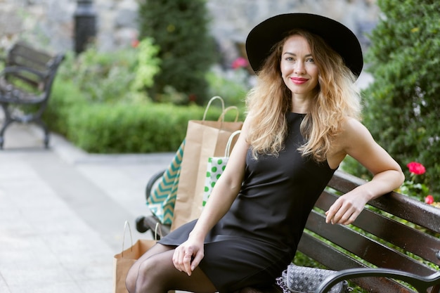 Nice young woman in a black felt hat and dress sits on a bench in a city park