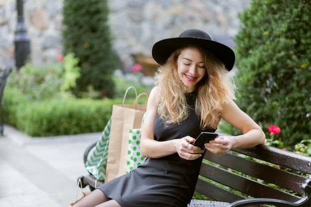 Nice young woman in a black felt hat and dress with shopping bag sits on a bench in city park and uses smartphone