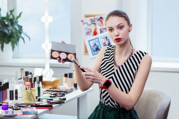 Nice young woman holding a powder palette while applying on the brush