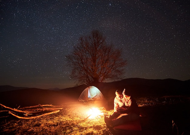 Night camping. Hikers resting near campfire under starry sky