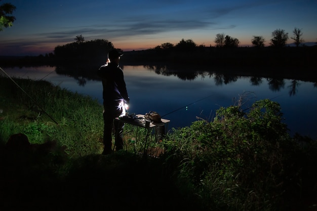 Night fishing, fisherman with a fishing rod standing on the shore of lake.