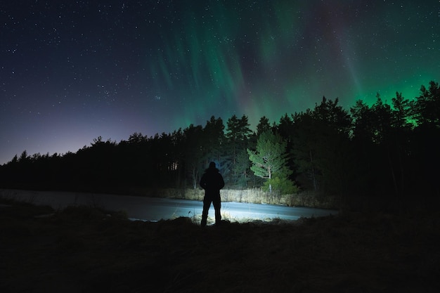 Night scene nature of Estonia Silhouette of a man with a flashlight in a dark forest with a starry sky and northern lights