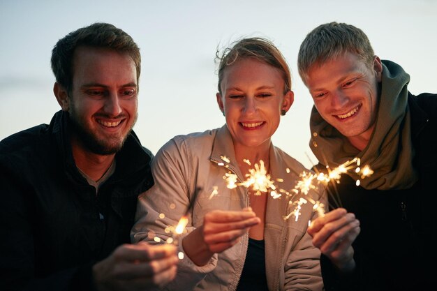 Photo night smile and sparklers with friends on beach together for holiday travel or vacation in summer celebration evening and fireworks with group of happy people outdoor on coast for new years