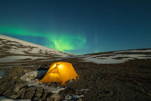 Night Starry Sky and Green Northern Lights over Glowing Yellow Tent in Khibiny Mountains at Winter Night. Russia.