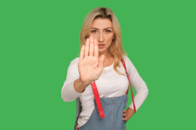 No access forbidden way Portrait of strict adult blond woman in stylish denim overalls showing stop block hand gesture rude rejection warning of danger studio shot isolated on green background