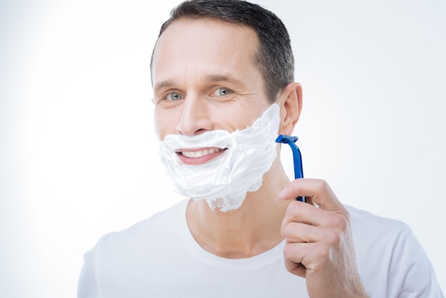 No beard. Joyful nice positive man holding a razor and looking at you while getting rid of his beard