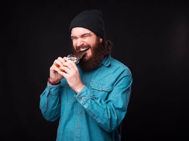 No diet. Bearded hipster man in blue shirt eating dark chocolate over