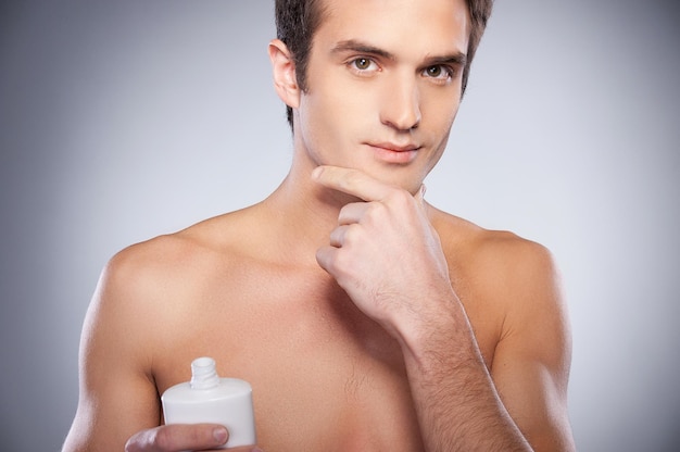 No irritation. Handsome young shirtless man applying cream at his face and looking at camera while standing against grey background