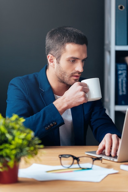 No time for break. Handsome young man in smart casual wear working on laptop and holding coffee cup while sitting at his working place in office