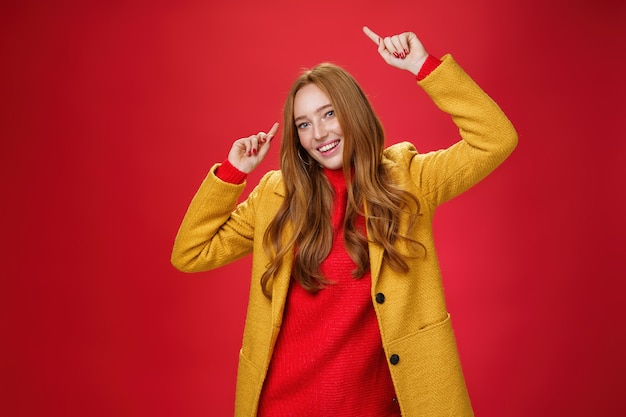 No time for sorrow dance and feel happy autumn came. Portrait of bright and cheerful charming redhead woman raising hands up moving to rhythm of music delighted posing in yellow coat over red wall.