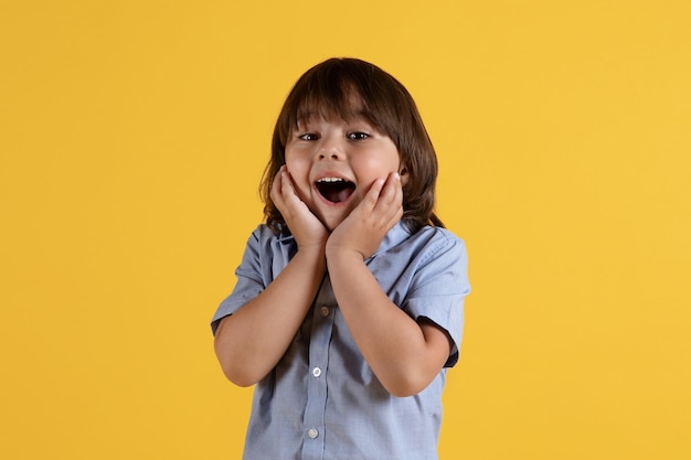 No way Surprised little boy touching his cheeks in amazement looking at camera with open mouth orange background