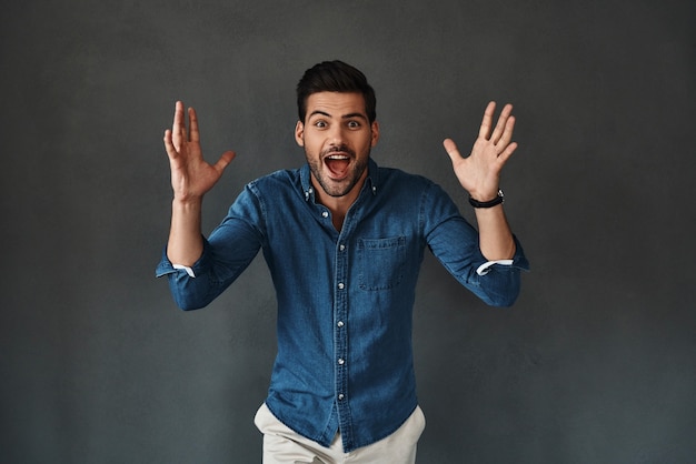 No way! Surprised young man in denim shirt looking at camera and keeping mouth open while standing against grey background