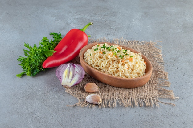 Noodle in a bowl on a burlap next to vegetables, on the marble surface.