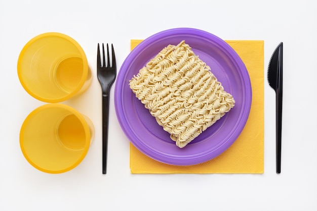 Noodles in a plastic lilac plate, yellow glass, black fork and knife, top view.