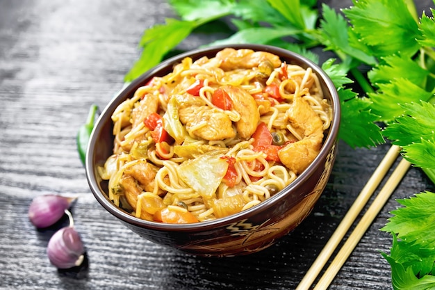 Noodles with cabbage, chicken breast, sweet red peppers, onions and peas, seasoned with soy sauce in a bowl, garlic and celery leaves on black wooden board background