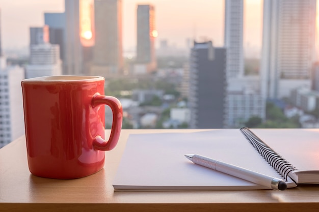 Note paper, book, pen and red coffee cup on wood table with building background