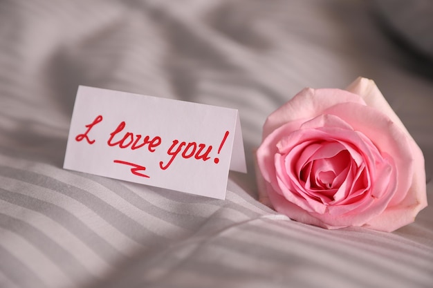 Note with handwritten text I Love You near pink rose on bed closeup Romantic message