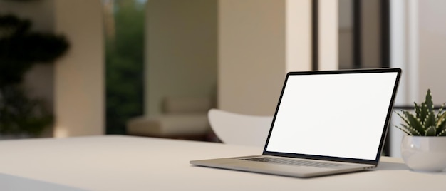 Notebook laptop white screen mockup is on white tabletop over blurred living room background