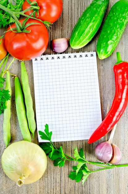 Notebook, red tomatoes, red hot pepper, parsley, garlic, onion, cucumber, green bean pods on an old wooden board