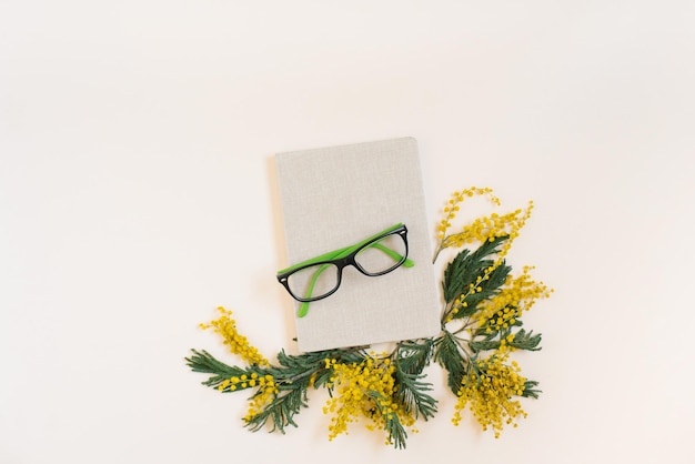 Notebook surrounded by yellow mimosa flowers on a light background with glasses Flat lay freelancer or blogger