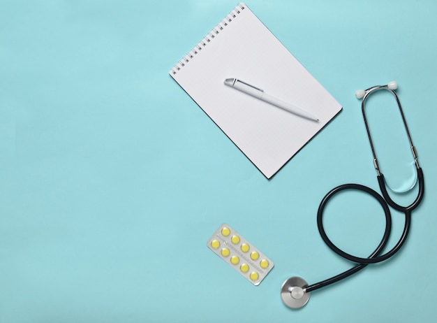 Notebook, tablets and medical stethoscope on a blue pastel background, minimalist trend, top view, medical workplace