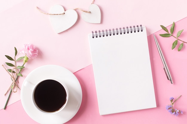 Notebook with clean page, cup of coffee, flower, pen and wooden heart on pink background. Top view, flat lay style.