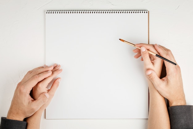 Notebook with empty sheet on a white wall with brush in hands, top view. Flat lay with copy space. Office concept