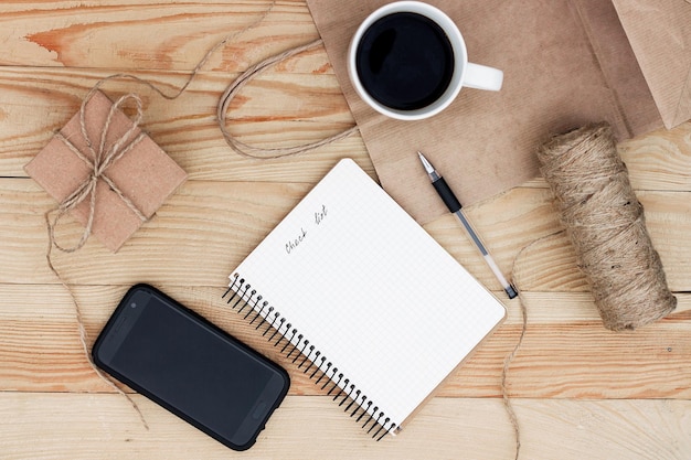 A notebook with an inscription Check list laying on wooden table. It is surrounded by phone, black pen, cup of coffee, present, rope twine and a paper bag.