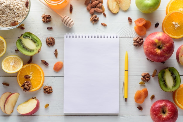 Notebook with pen surrounded by apples, kiwi, dried fruits, oranges and apples. The concept of a healthy diet and shopping list.