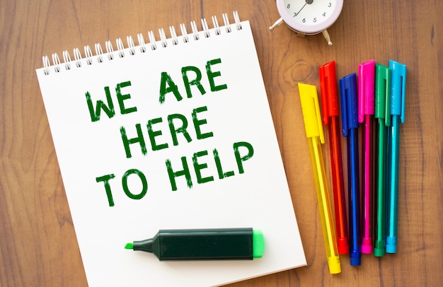 A notebook with the text WE ARE HERE TO HELP on a white sheet lies on a brown wooden table with colored pens. Business concept.