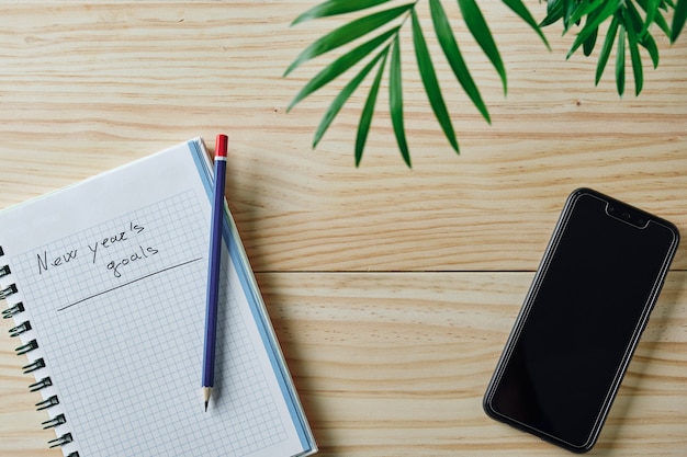 Notebook with the words new year's goals written on a natural wooden background with a blue and red pencil on top, some green leaves on top and a smartphone next to it