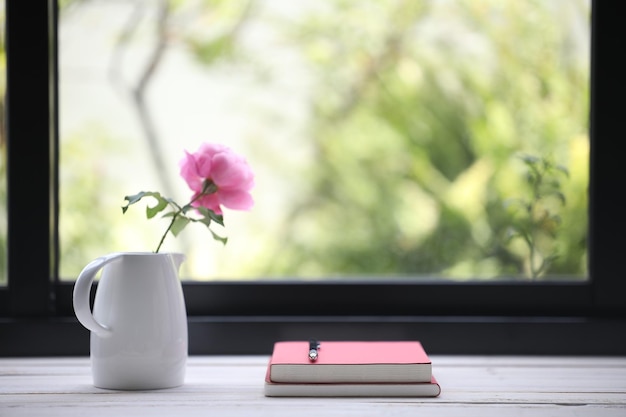 Notebooks and pink rose on wooden table in front of window