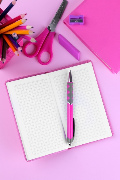 Notepad with a pen and other school supplies in pink  background.