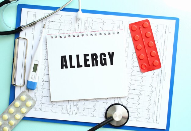 A notepad with the text ALLERGY lies on a blue medical clipboard with a stethoscope and pills on a blue background. Medical concept.