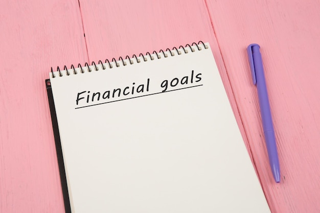 Notepad with text Financial goals and pen on pink wooden table