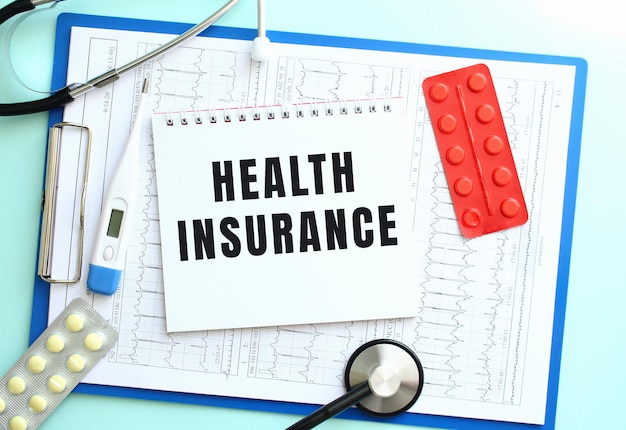 A notepad with the text health insurance lies on a medical clipboard with a stethoscope and pills on